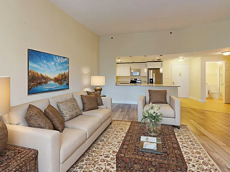 Elegant living room with cozy beige sofas, stylish decor, and open-plan view to a modern kitchen, featuring warm lighting and a serene artwork accent.