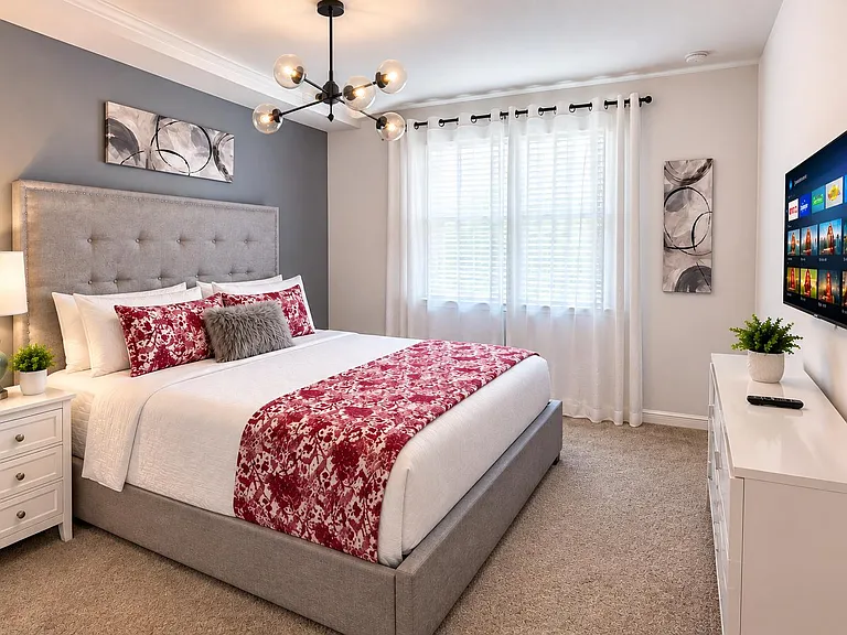 Spacious bedroom with a plush upholstered bed, nightstand lighting, large window with curtains, and a dresser with TV.