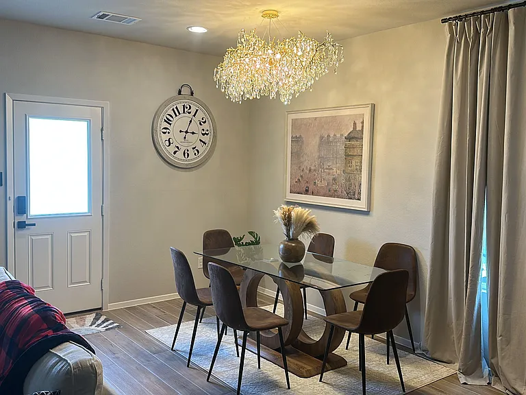 Dining area with a glass-top table for six, statement chandelier, and large window with full-length curtains.