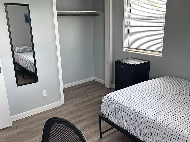 Private bedroom with closet storage, wood-look floors, and a window with blinds; includes a mini fridge and furnished setup.
