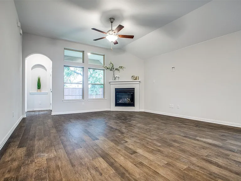 Spacious room featuring wood-finish flooring, a corner fireplace with white mantel, a ceiling fan, vaulted ceiling, and multiple windows