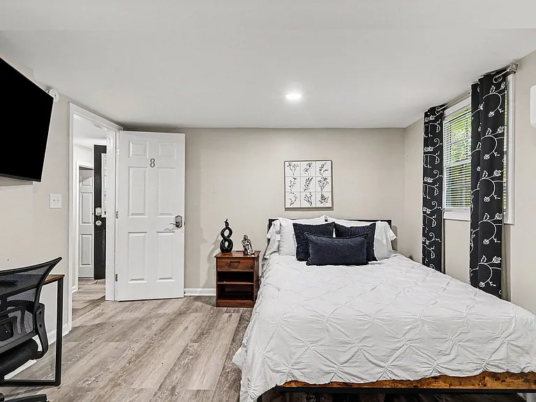 Furnished bedroom with a full bed, nightstand, desk seating, and wall-mounted TV, plus window coverings and AC.