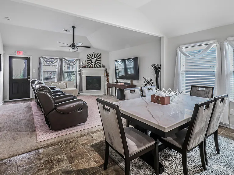 Open-concept living and dining area with large windows, ceiling fan, fireplace, and space for a full dining set.