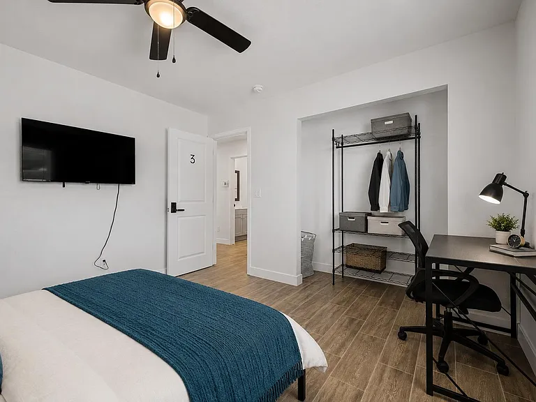 Private bedroom with bed, wall-mounted TV, work desk, and open closet storage with shelving for easy organization.