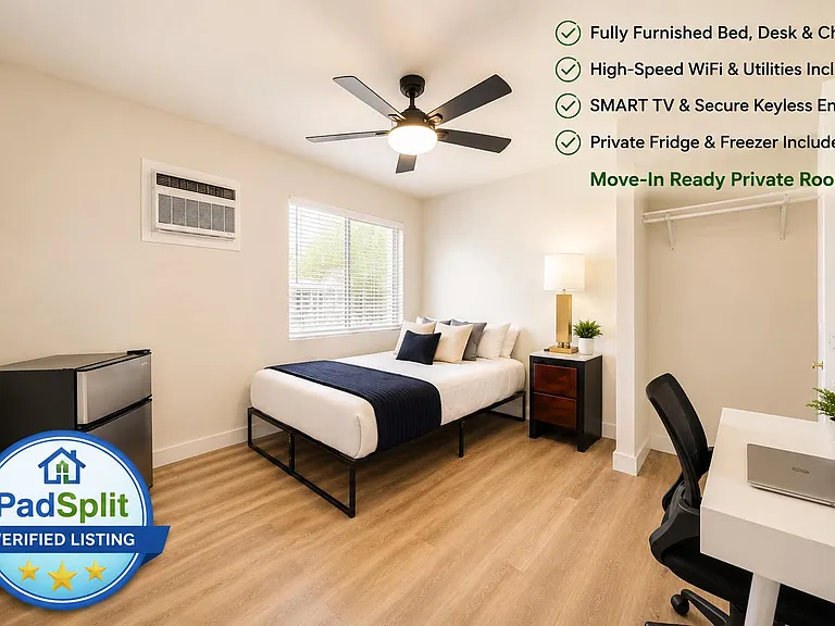 Furnished private bedroom with full bed, desk workspace, mini fridge, and open closet storage alongside a large window.