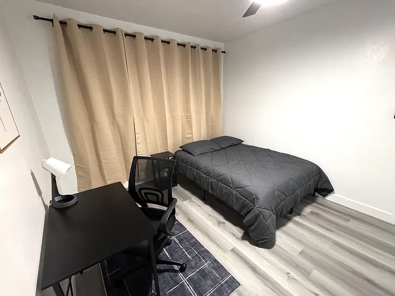 Furnished private bedroom with a comfortable bed, nightstand, and dedicated desk workspace, plus window coverings for privacy