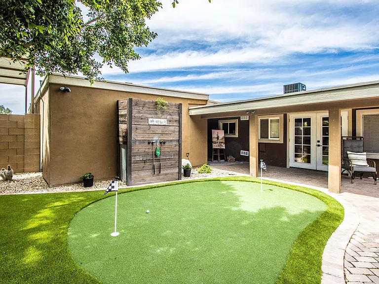 Private putting green at Casa Del Sol, Tempe rental home. Features a lush artificial turf green with golf flags, a rustic wood accent wall with putter storage, a covered patio, and desert landscaping. Fun backyard amenity for a top Tempe, AZ stay.