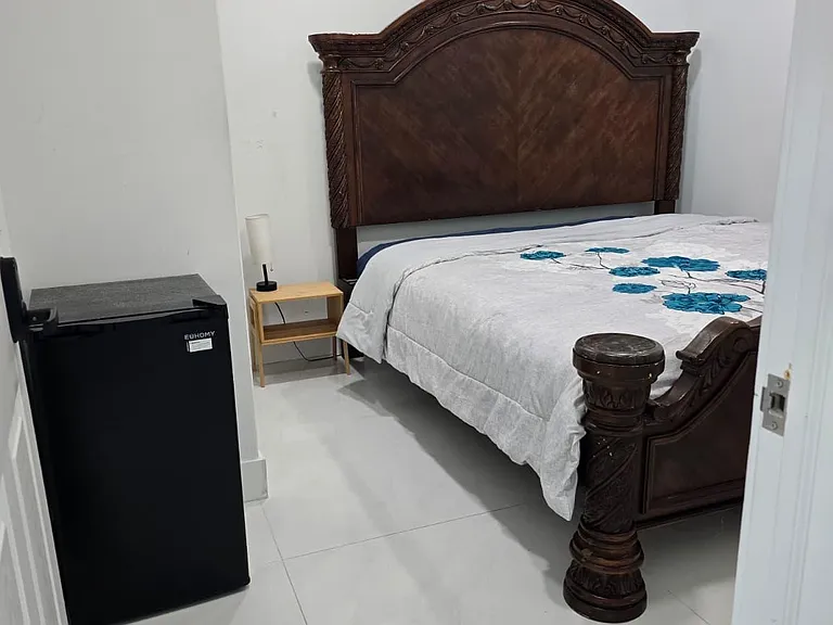 Private bedroom with a full bed, nightstand, and mini fridge, plus easy-care tile flooring and overhead lighting.