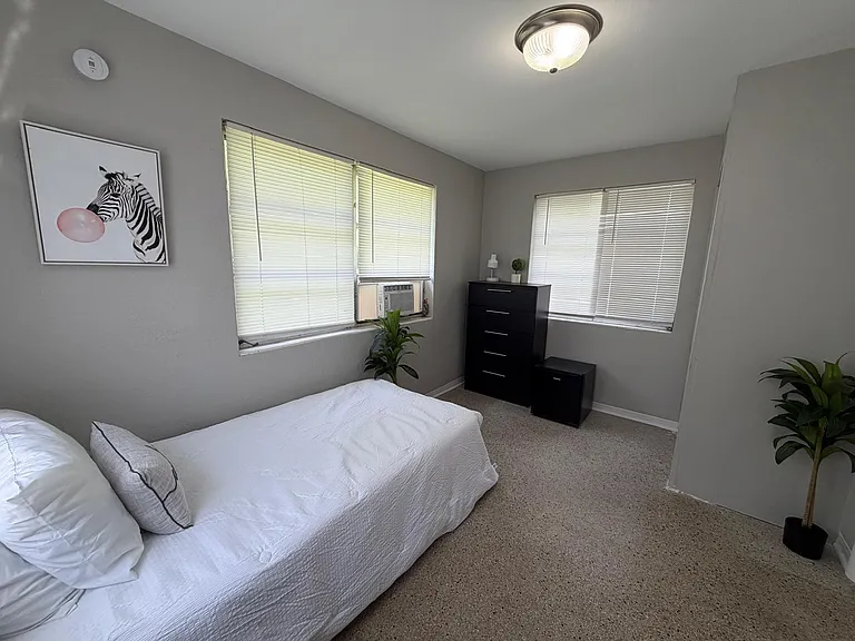Furnished bedroom with two windows, a dresser, and in-room AC, offering comfortable space and practical storage.