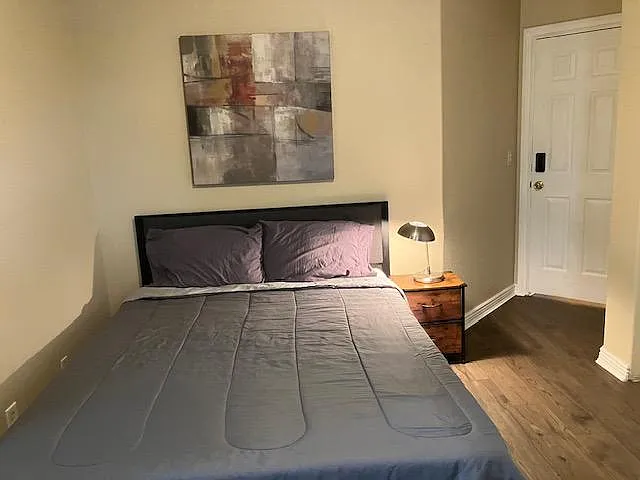 Bedroom features a queen-size bed, nightstand with lamp, and comfortable layout with neutral walls and durable flooring.