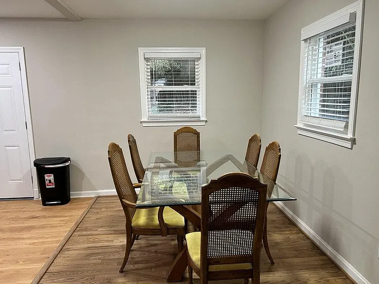 Dining area with a glass-top table for four, hardwood-style flooring, and two windows with blinds for natural light.