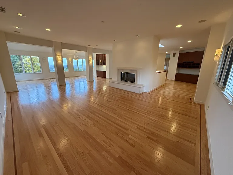 Grand living room and formal dining room featuring a fireplace, built-in buffet cabinetry, recessed lighting, rich wood flooring throughout and a wall of glass panels showcasing the bay view.