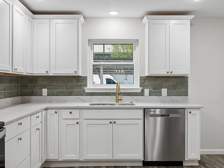 Kitchen featuring white shaker cabinetry with brass hardware, a white countertop, subway tile backsplash, stainless steel dishwasher, and a window above the sink