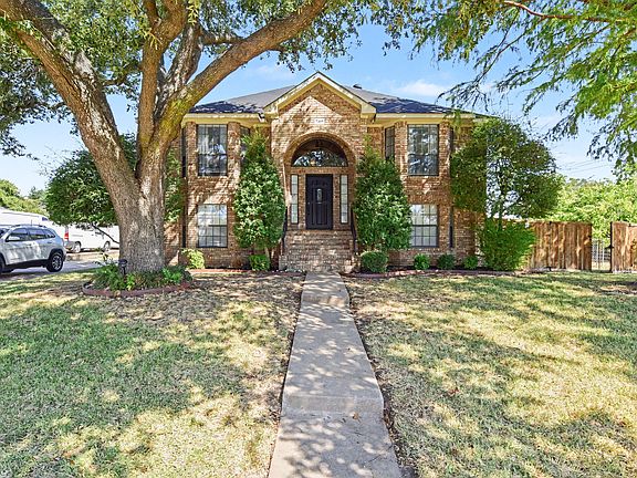 Brick two-story home with a defined front walkway, mature shade trees, and a landscaped entry with fenced side yard.