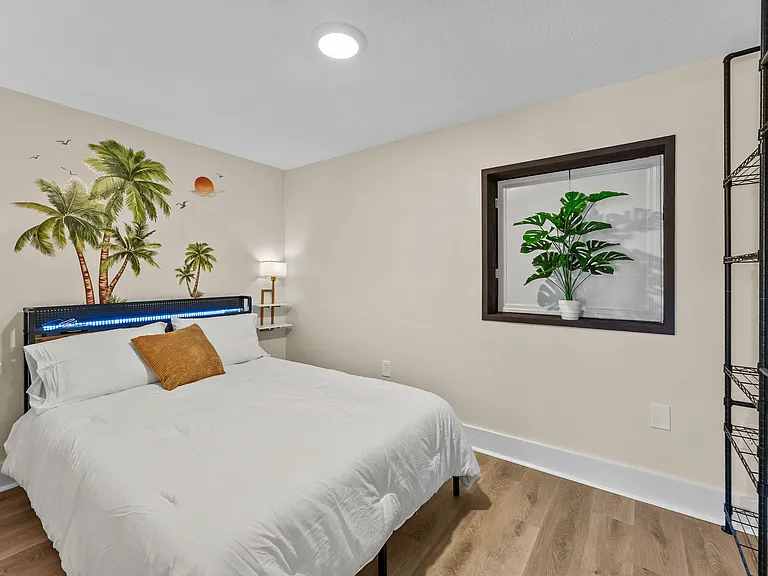 Furnished bedroom with a full-size bed, bedside lighting, and wood-style floors plus a window and open shelving for storage.