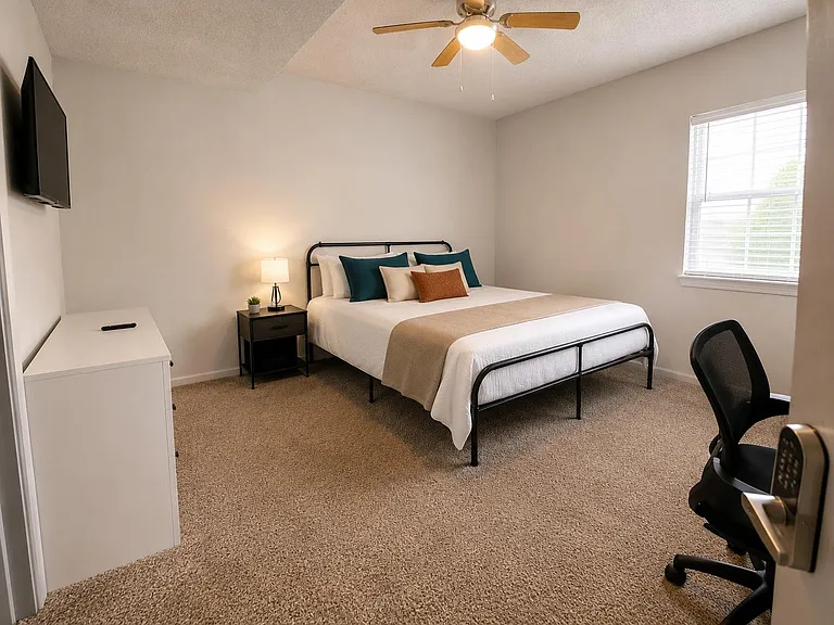 Furnished bedroom with a full bed, nightstand and lamp, desk chair, wall-mounted TV, and a large window with blinds.