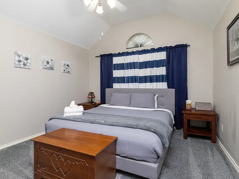 Bedroom 2 features a spacious king-size bed with soft gray bedding, bedside tables, room-darkening curtains, simple wall art, and a storage chest for convenience.