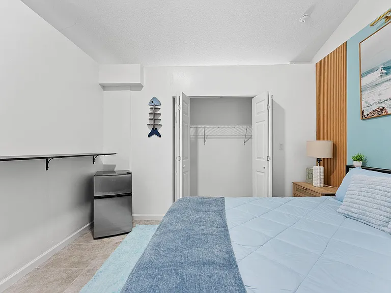 Furnished bedroom with a full bed, nightstand, mini fridge, and an open closet with shelving for easy storage.