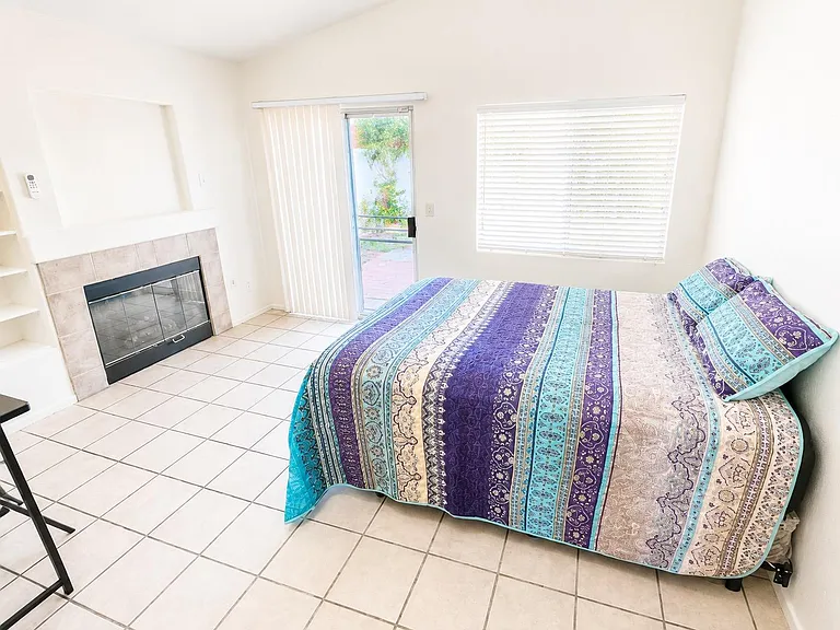 Spacious bedroom with tile flooring, a large window with blinds, and sliding door access to the patio; features a fireplace.