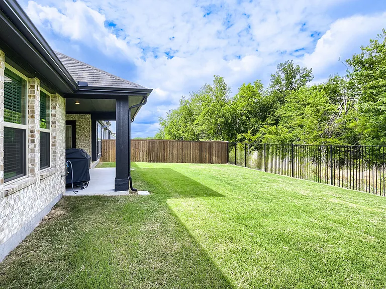 Expansive backyard featuring a manicured lawn, covered patio, two fencing types, and mature trees beyond the property line