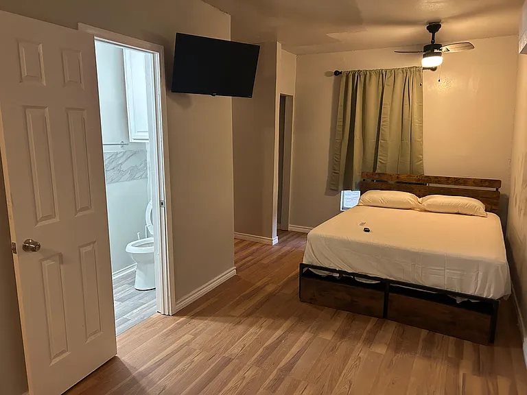 Furnished bedroom with wood-look floors, ceiling fan, wall-mounted TV, and convenient access to a nearby bathroom.