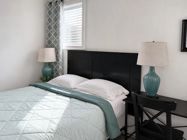 Furnished bedroom with a full bed, nightstands, and a dedicated desk area, plus a window with blinds and curtains.