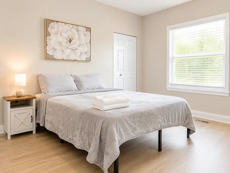 Comfortable bedroom with a full bed, bedside table and lamp, plus a large window with blinds for natural light.