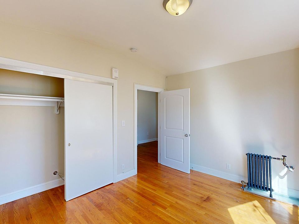 An empty room with wooden floors. Features include an open closet with sliding doors, a door to another room, a radiator, and natural light from a window, creating a warm atmosphere.