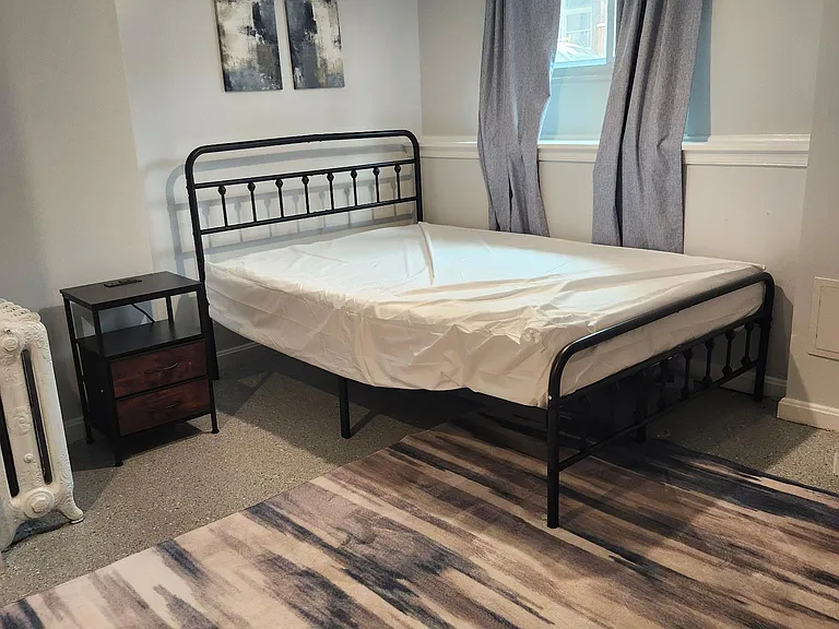 Furnished bedroom with metal bed frame, nightstand storage, and a window with curtains for natural light and privacy.