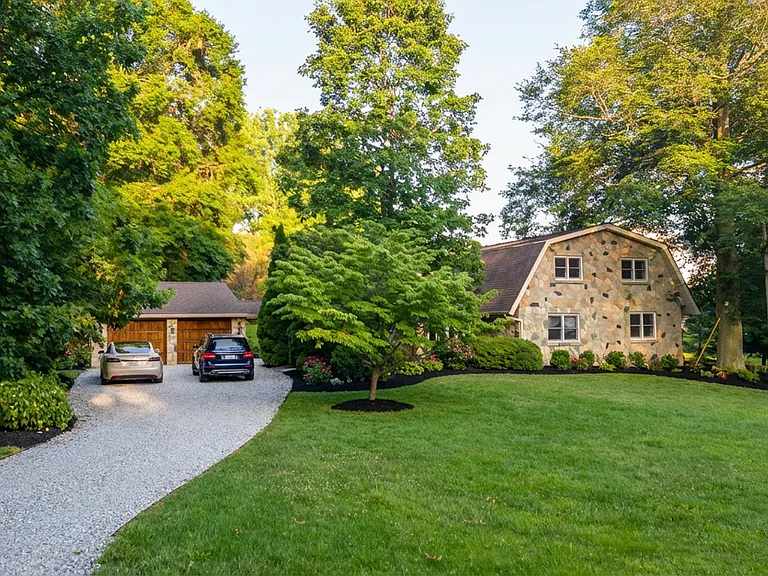 This stunning Dutch-style stone home at 4365 Centennial Lane in Ellicott City is surrounded by lush greenery and mature trees, creating a serene and private setting. A long gravel driveway leads to the inviting entrance, complemented by a manicured lawn.