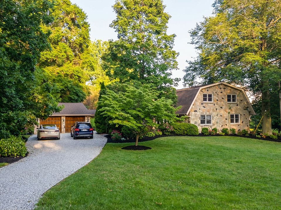 This stunning Dutch-style stone home at 4365 Centennial Lane in Ellicott City is surrounded by lush greenery and mature trees, creating a serene and private setting. A long gravel driveway leads to the inviting entrance, complemented by a manicured lawn.