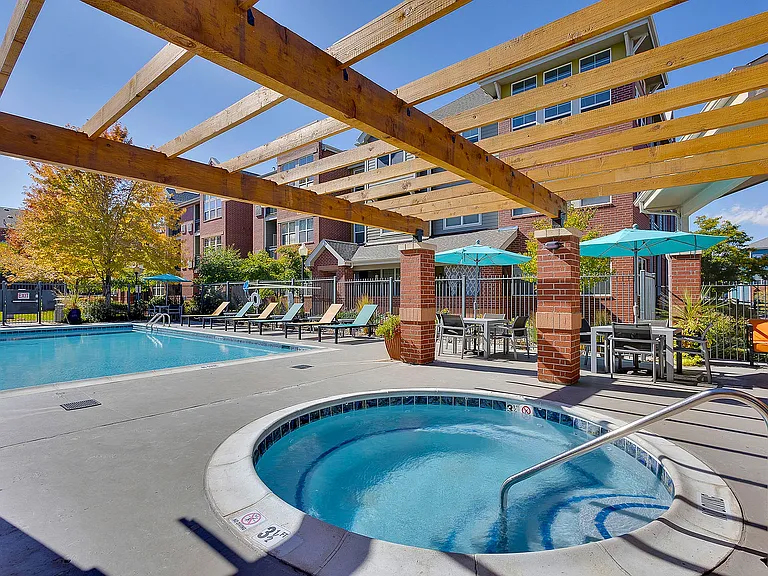 Inviting outdoor pool area with sun loungers, a hot tub, and stylish pergola, surrounded by vibrant landscaping and modern apartments.