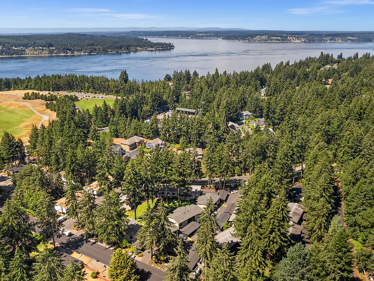 A scenic aerial view of the apartment community surrounded by mature evergreens, nearby waterfront views, golf course greenery, and Pacific Northwest scenery.