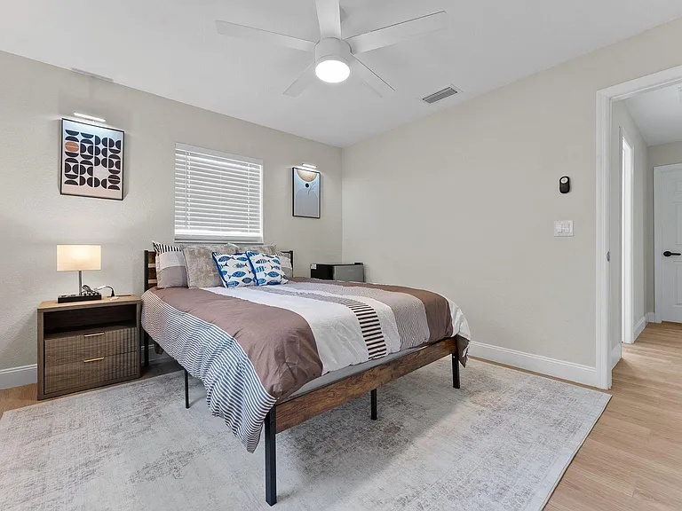Furnished bedroom with a queen bed, nightstand, ceiling fan, and window blinds, plus easy access to the hallway.
