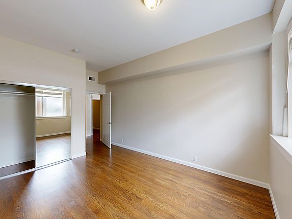 Empty room with wooden floors, featuring a large window with blinds on the right and a mirrored closet reflecting another window.