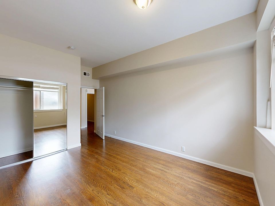 Empty room with wooden floors, featuring a large window with blinds on the right and a mirrored closet reflecting another window.