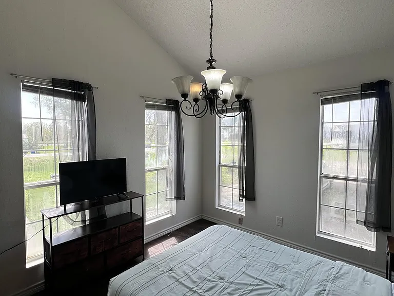 Spacious bedroom with three large windows, dark wood flooring, and an overhead chandelier for comfortable everyday living.