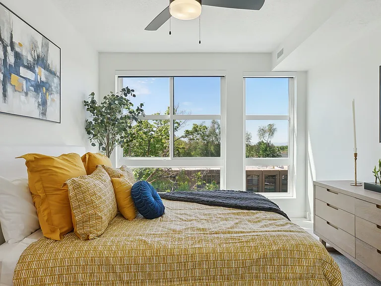 Liberty Duet Apartments, Salt Lake City, Utah; A bedroom with a bed, pillows, a ceiling fan, and a window overlooking a tree-filled area.