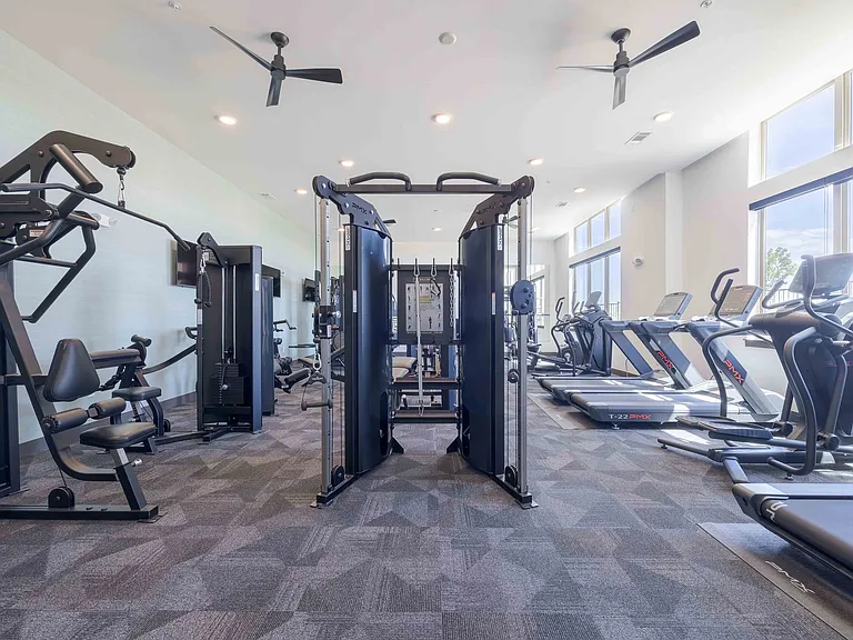 Cable functional trainers and leg machines anchor the strength training area of the Garrett Parker fitness center, with cardio equipment visible near the windows beyond.
