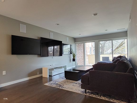 Furnished living room with couch, coffee table, TV console, & 3 large flat screen TVs.