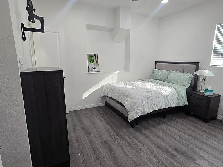 Furnished bedroom with a full/queen bed, nightstand, and wood-look flooring, plus a window with blinds for natural light.
