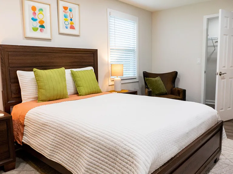 Furnished bedroom with a queen bed, nightstands, and reading chair, plus a window with blinds and an attached closet.