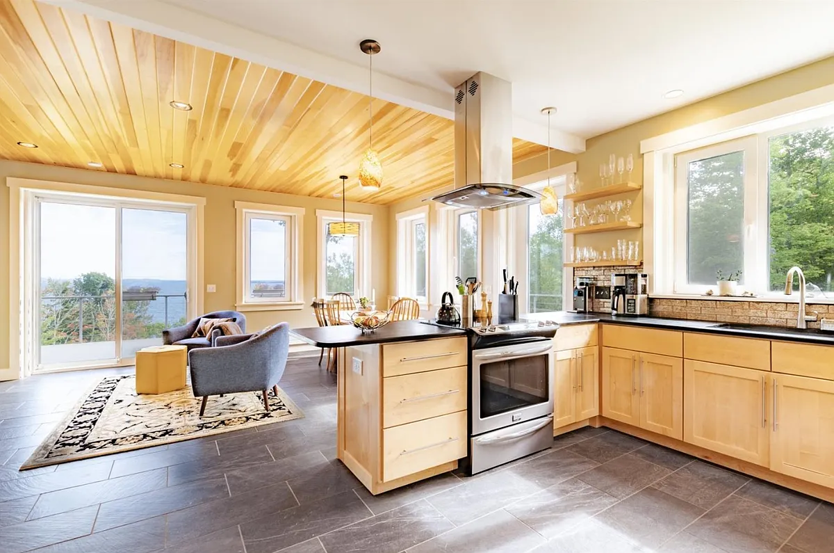 Kitchen area with dining and sitting area beyond. Modern appliances, custom cabinets, tile floor, open shelving and glowing wood ceiling. - Shore Rd