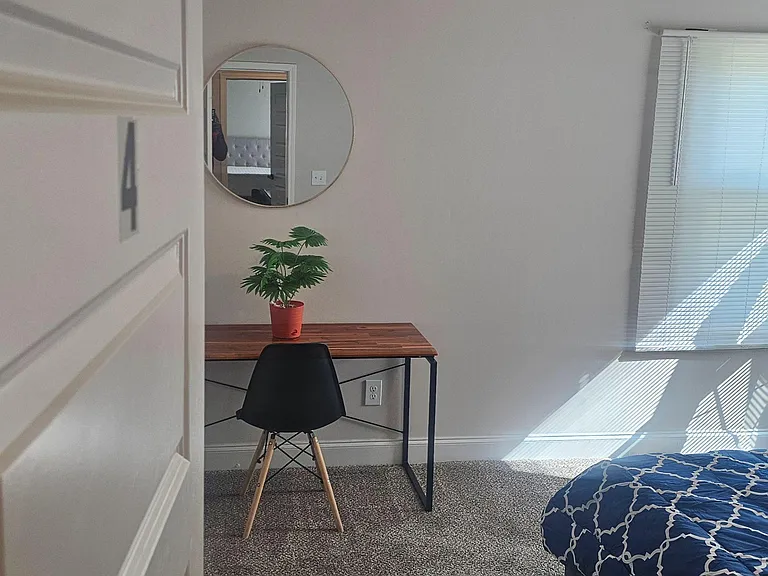 Carpeted bedroom with a work desk, chair, and window with blinds, offering a comfortable layout for rest and study.