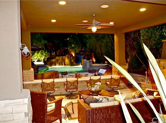 Outdoor covered room with lighting, audio speakers, stone fireplace, large travertine bar overlooking pool and kitchen area.