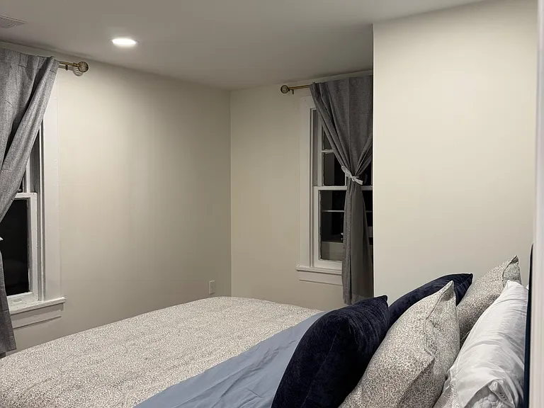 Furnished bedroom with a full-size bed, neutral walls, and two windows with curtain panels for ventilation and daylight.