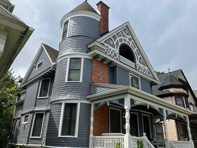 Exterior showing Victorian details including turret, chimney, mosaic surrounding 3rd floor balcony, and front porch with filigree and white wicker patio furniture.