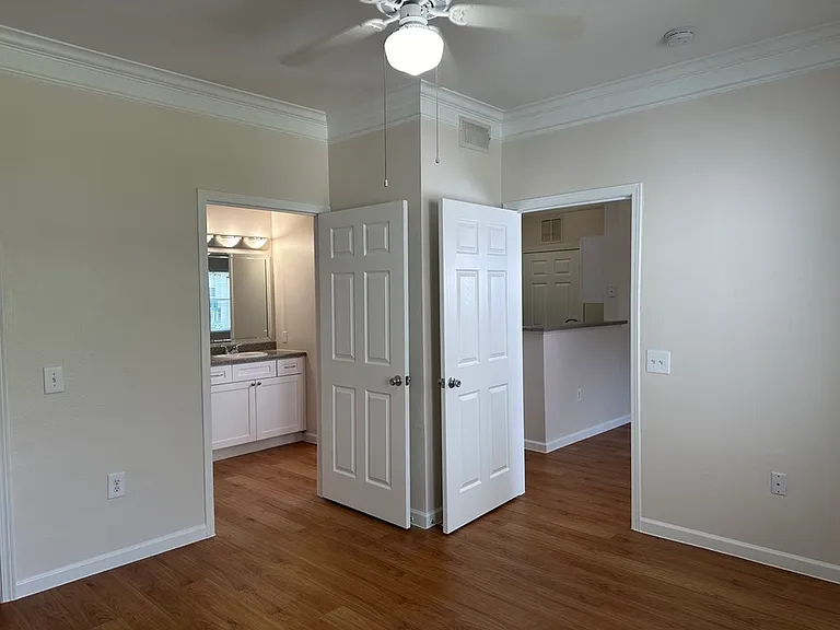 Bright room with wood-style floors, featuring elegant crown molding, a ceiling fan, and doorway views into a modern bathroom and kitchen. Bright room with wood-style floors, featuring elegant crown molding, a ceiling fan, and doorway views into a modern b