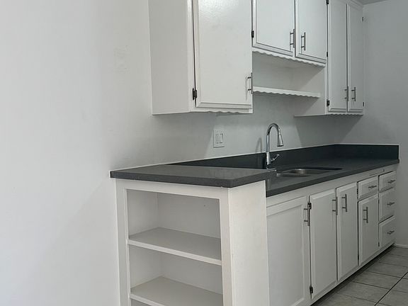 White kitchen cabinets with gray quartz countertops with chrome goose neck faucet. White open built in shelving and tan/beige flooring.