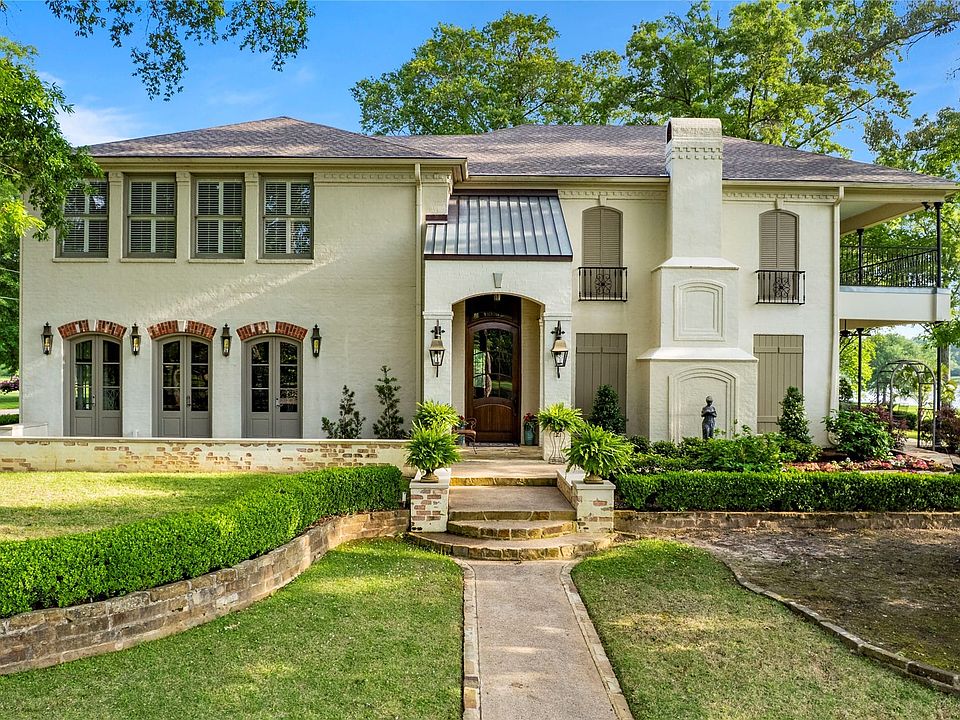 Stucco exterior with a metal awning over the entryway, featuring an upper-level balcony, ground-floor arched French doors, and mature trees